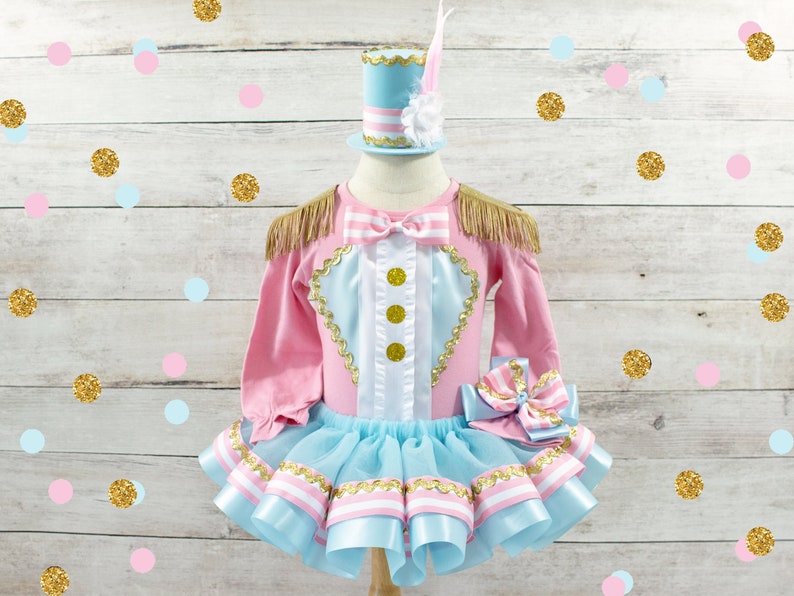 May include: A pink and blue circus-themed outfit with gold accents. The long-sleeved top has gold fringe epaulets and gold buttons. The tutu skirt is light blue with pink and white ribbon trim. A matching hat with a feather is included.