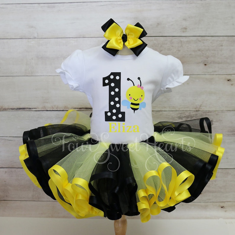 May include: A white t-shirt with a black polka-dotted number 1 and a cartoon bee graphic, paired with a black and yellow tutu skirt and a matching bow. The name "Eliza" is printed in yellow. The outfit is designed for a first birthday.