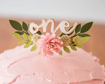 Könnte beinhalten: Ein rosa gefrosteter Kuchen mit einem weißen Papier-Topper, auf dem "one" steht, mit grünen Blättern und rosa Papierblumen.