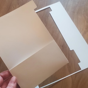 Puede incluir: Una carpeta blanca con un dise&ntilde;o recortado, que cubre parcialmente una carpeta beige.