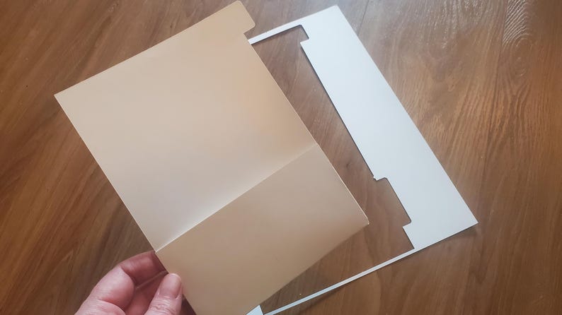 Puede incluir: Una carpeta blanca con un dise&ntilde;o recortado, que cubre parcialmente una carpeta beige.
