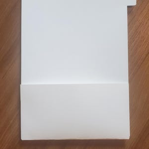 Puede incluir: Una carpeta de archivos blanca con una pesta&ntilde;a en la esquina superior derecha. La carpeta est&aacute; doblada por la mitad para crear un bolsillo.