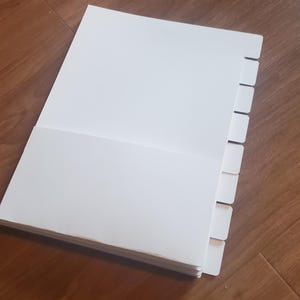 Puede incluir: Un cuaderno blanco de m&uacute;ltiples p&aacute;ginas con un juego de divisores a lo largo del borde derecho. Los divisores son blancos y tienen un dise&ntilde;o con pesta&ntilde;as.