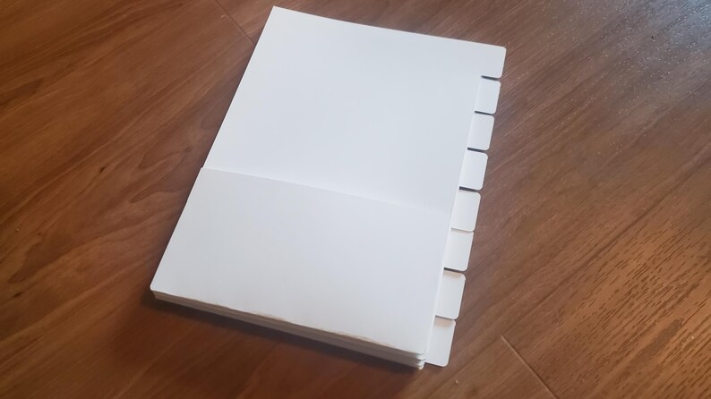 Puede incluir: Un cuaderno blanco de m&uacute;ltiples p&aacute;ginas con un juego de divisores a lo largo del borde derecho. Los divisores son blancos y tienen un dise&ntilde;o con pesta&ntilde;as.