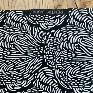 May include: Black and white fabric with a repeating pattern of abstract swirls and lines. The fabric has a label that reads "PENTIK JAKALA".