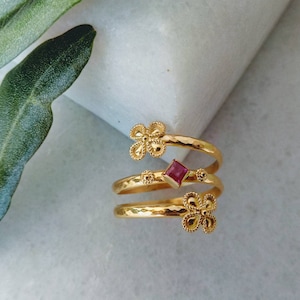 May include: Gold ring with three bands, each featuring a delicate flower design. The center band has a small square red gemstone.