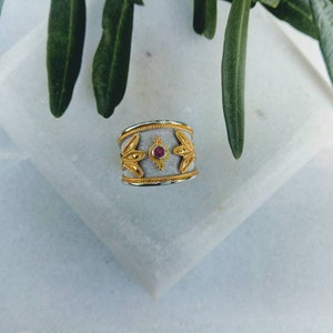May include: A silver and gold ring with a red gemstone set in the center. The ring has a decorative design with floral motifs.