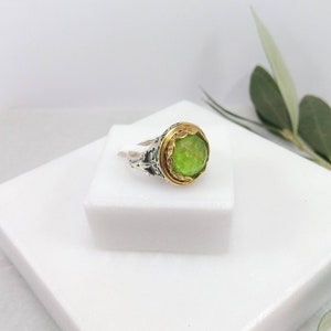 Può includere: Un anello d'argento con una pietra preziosa verde incastonata in una ghiera d'oro. L'anello presenta un design intricato sulla fascia.
