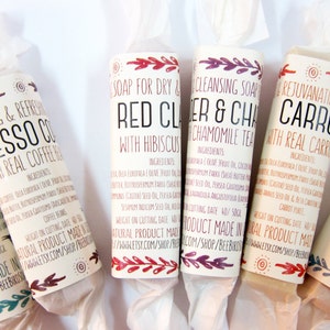 Puede incluir: Seis barras de jabón natural diferentes envueltas en papel blanco. Los jabones están etiquetados con nombres e ingredientes, que incluyen "Arcilla roja con hibisco", "Lavanda y té de manzanilla", "Zanahoria rejuvenecedora con zanahoria real" y "Sólido con arcilla blanca".