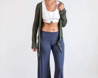 May include: An olive green long-sleeve open cardigan, a white scoop-neck crop top, and slate blue high-waisted wide-leg pants. A long necklace with a dark, textured pendant hangs down the center of the chest, completing the layered outfit.