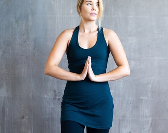 May include: A woman wearing a teal tank top and black leggings, standing with hands together in a prayer position. The outfit is suitable for yoga or other fitness activities. The background is a gray concrete wall.