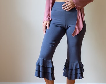 May include: A woman is wearing a white tank top with a long, beaded necklace, a dusty rose cardigan, and dark gray cropped pants with a ruffled hem.