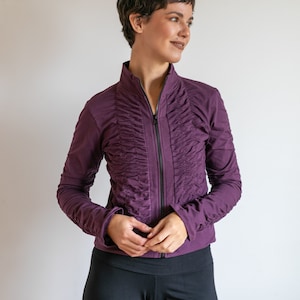 May include: A woman wearing a purple zip-up jacket with a ruffled front and black pants.