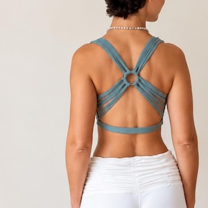 Puede incluir: Un top corto de color verde azulado con diseño de tiras en la espalda y un anillo central, combinado con pantalones blancos de pierna ancha con una banda fruncida de cintura alta. El conjunto se muestra de espaldas, destacando las tiras.