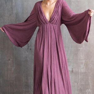 May include: A floor-length, deep mauve maxi dress featuring a deep V-neckline with gathered fabric detailing at the bodice and wide, flowing bell sleeves. The garment has a fitted waistline that transitions into a loose, floor-sweeping skirt.
