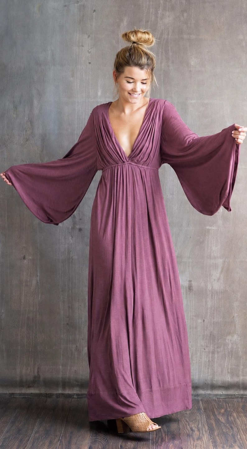 May include: A floor-length, deep mauve maxi dress featuring a deep V-neckline with gathered fabric detailing at the bodice and wide, flowing bell sleeves. The garment has a fitted waistline that transitions into a loose, floor-sweeping skirt.
