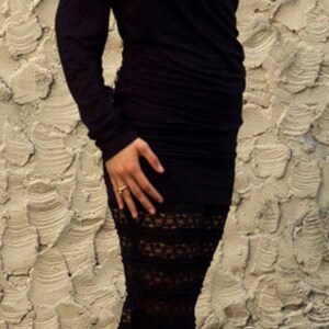 May include: A woman wearing a black long-sleeved dress with a lace bottom and black heeled sandals. She is standing in front of a textured wall.