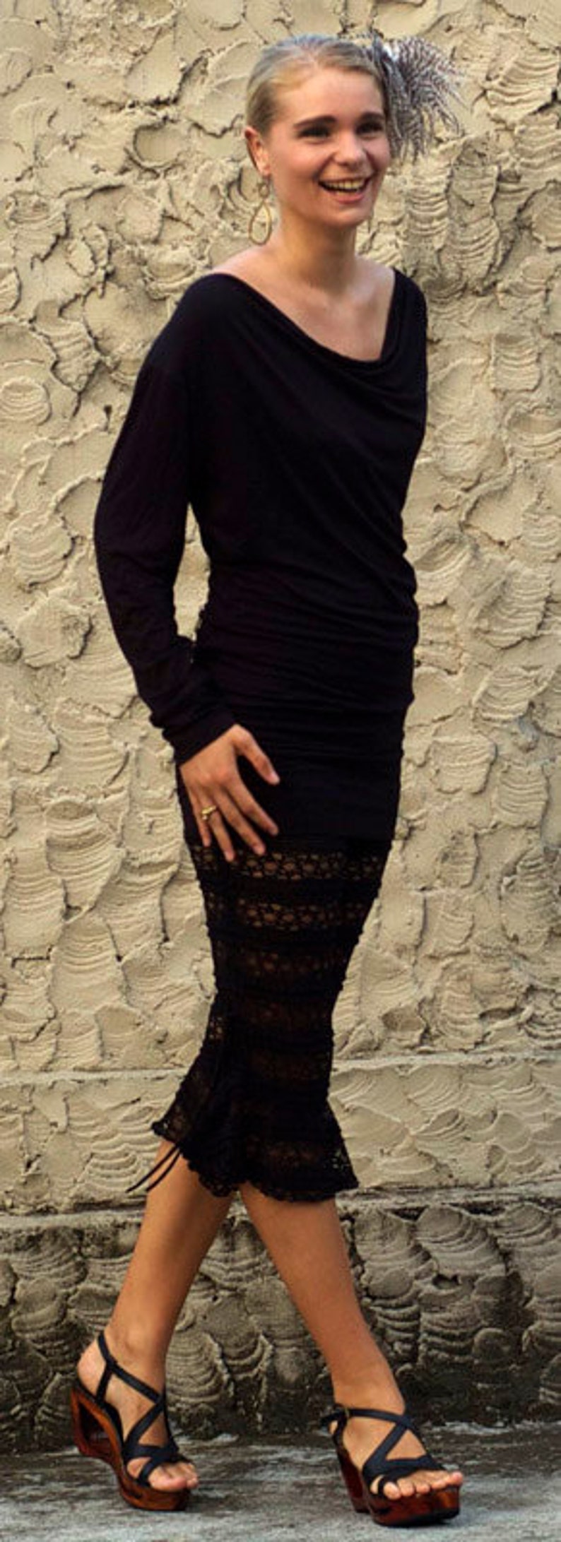 May include: A woman wearing a black long-sleeved dress with a lace bottom and black heeled sandals. She is standing in front of a textured wall.