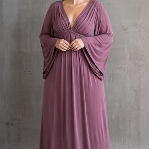 May include: A floor-length mauve maxi dress featuring a deep V-neckline with gathered fabric detailing at the bodice and wide, flowing bell sleeves. The garment has a fitted waistline that transitions into a loose, floor-sweeping skirt.