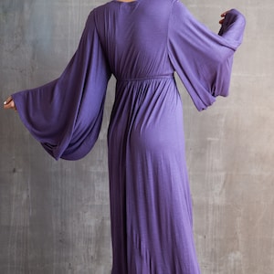 May include: A floor-length purple dress featuring long, wide bell sleeves and a gathered waistline. The garment has a V-neckline and a flowing, loose-fitting skirt that extends to the floor, crafted from a soft, solid-colored fabric.