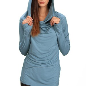 May include: A woman wearing a light blue hooded top with a cowl neck. The top is made of a soft, stretchy fabric. The woman is wearing black leggings with a patterned design.