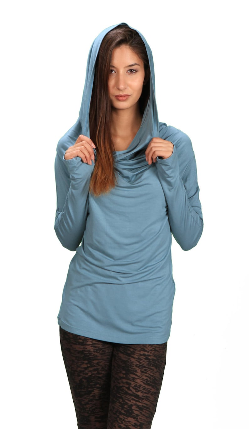 May include: A woman wearing a light blue hooded top with a cowl neck. The top is made of a soft, stretchy fabric. The woman is wearing black leggings with a patterned design.