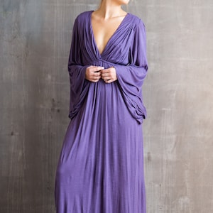 May include: A floor-length purple dress featuring a deep V-neckline, gathered fabric at the bodice, and long, loose-fitting sleeves that drape from the shoulders. The garment has a flowing skirt and a solid, textured purple fabric throughout.