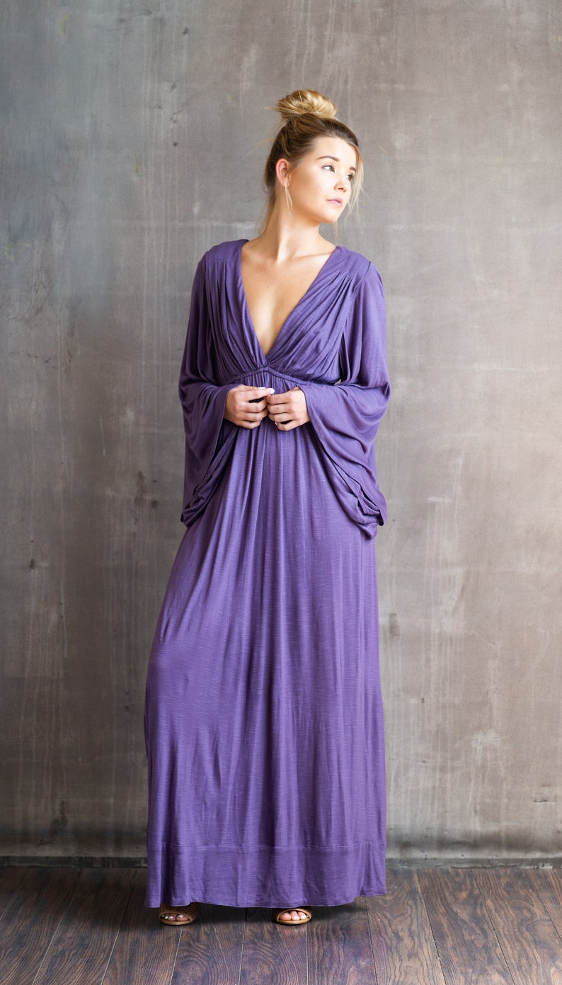 May include: A floor-length purple dress featuring a deep V-neckline, gathered fabric at the bodice, and long, loose-fitting sleeves that drape from the shoulders. The garment has a flowing skirt and a solid, textured purple fabric throughout.