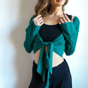 Op de afbeelding: Een vrouw draagt een groene, langarmige wikkeltop die in de taille is geknoopt over een zwart tanktop en een zwarte broek.