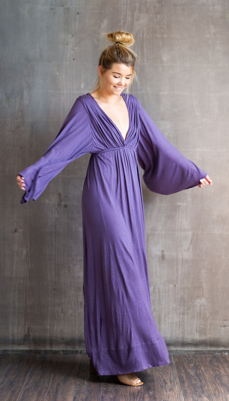 May include: A floor-length purple maxi dress featuring a deep V-neckline, an empire waist, and long, wide bell sleeves. The garment is crafted from a solid-colored, textured fabric that flows downward from the gathered bodice to the hemline.