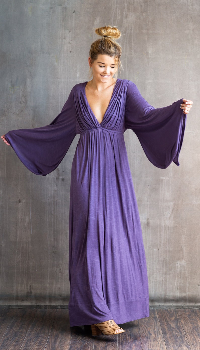 May include: A floor-length purple maxi dress featuring a deep V-neckline, gathered fabric at the bodice, and wide, flowing bell sleeves. The garment has a fitted waistline that transitions into a loose, floor-sweeping skirt with a solid hem.