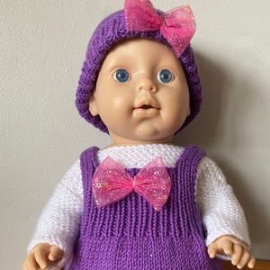 May include: A purple knitted doll dress with a white long-sleeved shirt underneath. The dress has a pink bow with glitter on the front and the doll is wearing a purple knitted hat with a pink bow on top. The doll is also wearing purple knitted booties with pink bows.