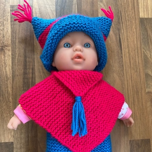Könnte beinhalten: Ein gestricktes Puppenoutfit in Pink, Blau und Gelb. Das Outfit beinhaltet eine Mütze, einen Poncho und eine Hose.