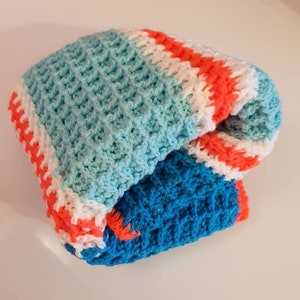 May include: A folded, handmade crochet blanket featuring a waffle stitch pattern. The blanket showcases a color scheme of light blue, white, orange, and dark blue. The edges are neatly finished, highlighting the craftsmanship of the item.