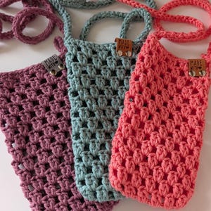 May include: Three handmade crochet phone bags in shades of pink, teal, and mauve. Each bag features a woven pattern, a long strap, and a small leather tag that reads "MADE WITH". The bags are designed to be worn as a crossbody.