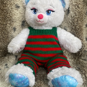 Può includere: Orso di peluche bianco con un vestito a righe verdi e rosse di maglia. L'orso ha gli occhi blu e un naso rosa. I piedi dell'orso hanno un tessuto blu con disegni di fiocchi di neve.