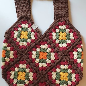 May include: A brown crocheted tote bag with a granny square pattern featuring red, white, green, orange, and yellow yarn. The bag has a brown crocheted strap.