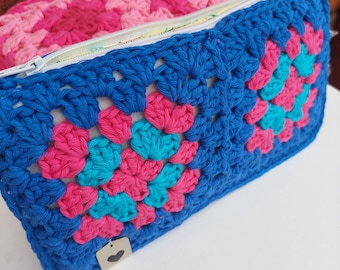 May include: A handmade crochet pouch in a granny square pattern. The pouch features a vibrant color scheme of blue, pink, and teal. The zipper is white, and the interior is lined with a light yellow fabric. A small tag with a heart is attached.