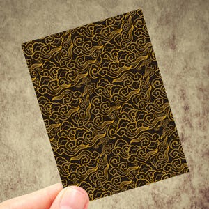 May include: A rectangular card with a repeating pattern of gold swirling cloud-like shapes on a dark brown background. The card is held by a hand against a textured, neutral-toned surface.