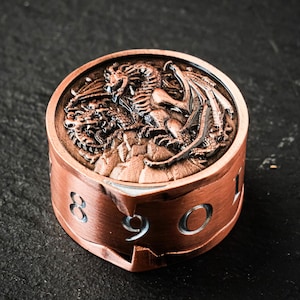 Puede incluir: Un contador de dragón D99 de color cobre con un relieve detallado de dos dragones. El contador tiene los números 8, 9 y 0 recortados en el lateral. El texto "D99 Dragon Counter" está en el lado izquierdo.