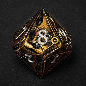 May include: A metallic gold and bronze polyhedral die with silver numbering. The die features intricate gear and clockwork-inspired detailing. The number 8 is prominently displayed on one face. The die is designed for tabletop gaming.