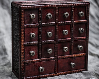 May include: A dark brown wooden drawer cabinet with 16 small drawers and 2 larger drawers. Each drawer has a silver knob. The cabinet has a decorative carved design on the front.