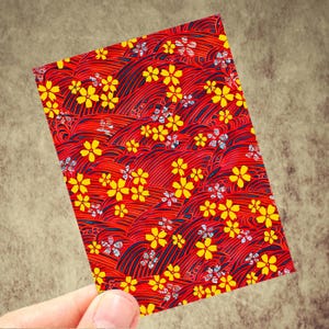 May include: A rectangular card with a vibrant red background featuring a repeating pattern of yellow flowers and stylized wave-like lines. The card is held by a hand, showcasing the intricate design and color contrast.