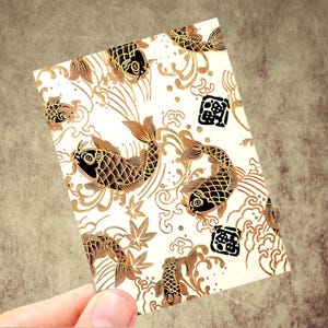 May include: A rectangular card featuring a repeating pattern of stylized koi fish in shades of brown and black against a cream background. The design includes water motifs and Japanese characters. The card is held by a hand.