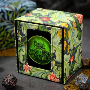 May include: A square dice box with a floral pattern in green, red, and yellow. The box has a window displaying a green, glowing image. Several dice are scattered around the box, including a black and red die.