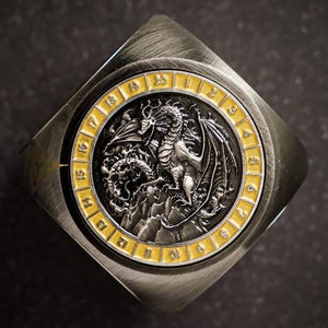 Puede incluir: Una torre de dados de metal plateado con un diseño de dragón y números dorados del 1 al 20 alrededor del borde.