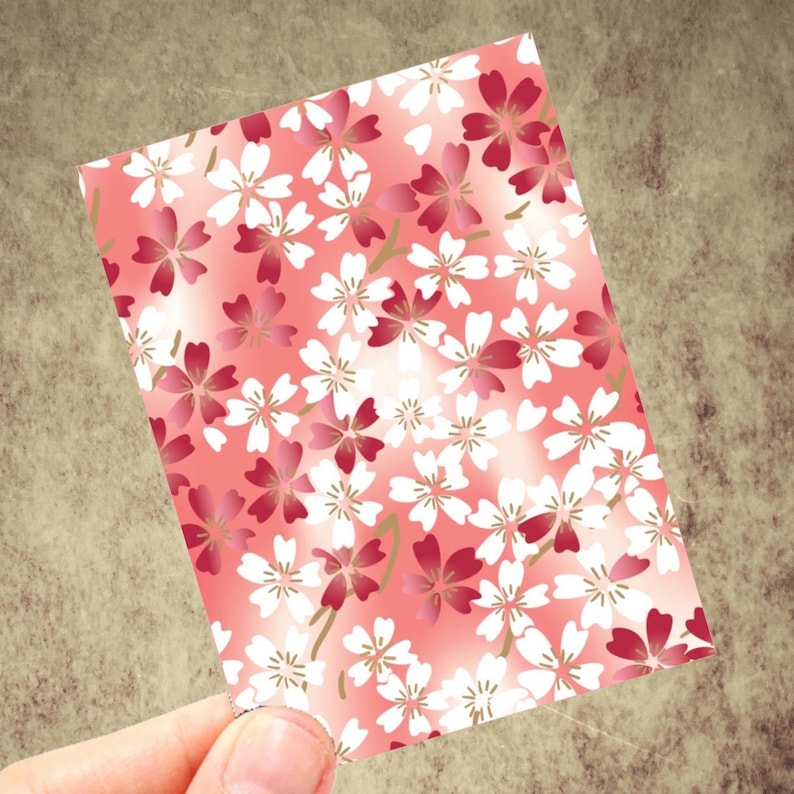 May include: A rectangular card featuring a repeating floral pattern of white and deep pink cherry blossoms on a gradient pink background. The card is held by a hand, showcasing the design's details.