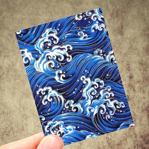 May include: A rectangular card featuring a stylized ocean wave pattern. The design showcases deep blue waves with white crests and gold accents. The card is held against a neutral background, highlighting the vibrant colors and intricate details of the wave illustration.