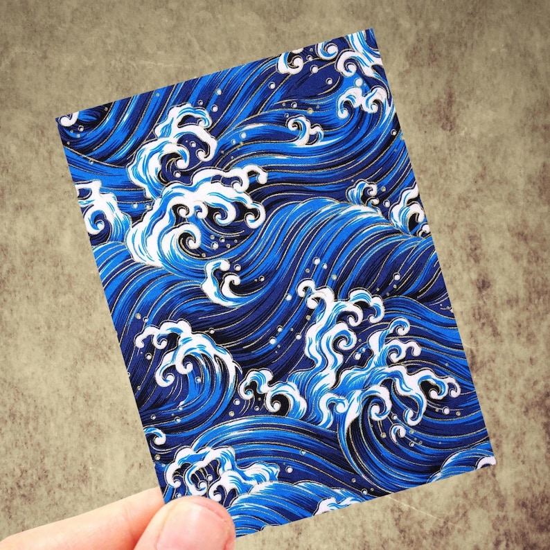 May include: A rectangular card featuring a stylized ocean wave pattern. The design showcases deep blue waves with white crests and gold accents. The card is held against a neutral background, highlighting the vibrant colors and intricate details of the wave illustration.