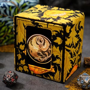 May include: A square deck box featuring a vibrant yellow and black floral pattern with a central circular emblem depicting a golden phoenix. The box is constructed with stitched edges and is surrounded by several multi-sided gaming dice.
